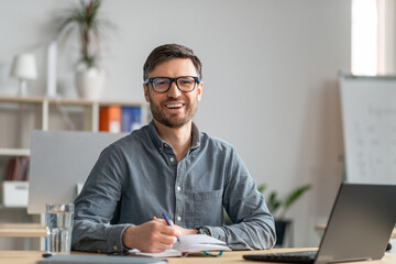 Poster - Happy mature entrepreneur having online training in office, using laptop, taking notes and smiling at camera, copy space