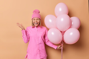 Canvas Print - Optimistic glad young woman wears pink hat and shirt laughs positively keeps palm raised holds bunch of inflated balloons enjoys festive occasion isolated over brown background. Holiday concept