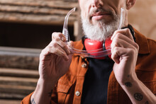 cropped view of bearded woodworker holding googles in workshop.