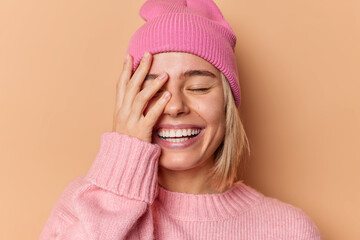 Wall Mural - Happy woman makes face palm smiles broadly shows white teeth closes eyes from satisfaction feels very glad hears excellent news wears hat and sweater isolated over brown background. Emotions concept