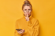 Discontent Serious Young Woman Demands Silence Keeps Index Finger Over Lips Asks To Be Quiet Frowns Face Holds Mobile Phone Wears Round Spectacles Casual Jumper Isolated Over Yellow Background