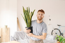 Happy Young Man Holding Plant In New Apartment Looking At Camera