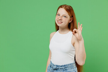 Little redhead kid satisfied happy smiling girl 12-13 years old wearing white tank shirt showing okay ok gesture isolated on plain green color background studio portrait. Childhood lifestyle concept.