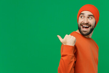 Wall Mural - Side view young smiling happy fun man 20s wear orange sweatshirt hat point thumb finger aside on workspace area mock up isolated on plain green background studio portrait. People lifestyle concept