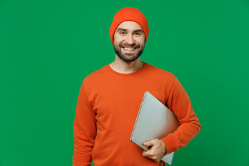 Wall Mural - Young smiling programmer student cheerful happy man 20s in orange sweatshirt hat hold closed laptop pc computer look camera isolated on plain green background studio portrait People lifestyle concept