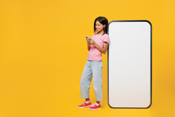 Full body little kid girl of African American ethnicity 12-13 years old in pink t-shirt near big huge blank screen mobile cell phone workspace copy space mockup isolated on plain yellow background