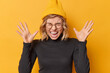 Photo of mad emotional woman yells keeps palms raised feels very angry quarrels with someone wears casual turtleneck hat and round spectacles isolated over yellow background. Emotions concept