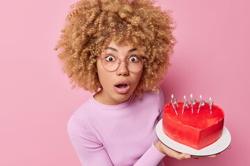 Wall Mural - Surprised shocked woman with curly bushy hair holds heart shaped delicious cake invites you on her birthday party hears something amazing wears spectacles casual jumper isolated on pink wall