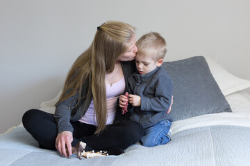 Cute three-year old blond boy sitting on bed dressed in casual clothes playing with animal toys while his mother gives him a kiss