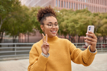 Wall Mural - pretty cheerful millennial girl wears round spectacles and yellow knitted sweater makes peace sign p