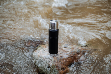 Wall Mural - A vacuum thermos shot close-up stands on a rock in the river. The concept of a thermo coffee cup, trekking in the spring in the mountains.