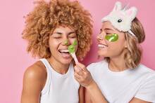 Cheerful Girlfriends Put On Green Facial Mask And Beauty Patches Take Care Of Skin In Domestic Conditions Smile Happily Wear Sleepmask Casual White T Shirt Pose Together Indoor Against Pink Wall