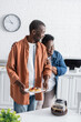 © LIGHTFIELD STUDIOS - happy african american woman hugging husband holding pancakes in kitchen.