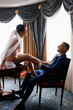 Side view of attractive brunette woman, wearing long veil, sexual lingerie and bridal shoes, sitting on table and touching bridegroom during wedding morning. 