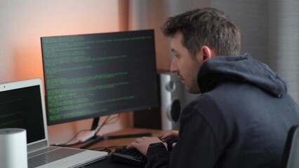 A hacker in a hood types program code. Young man criminal wearing black hoodie, typing on keyboard. Hacker attack.