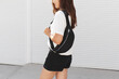 Woman, wearing white t-shirt, black shorts and fanny pack or waist pack, standing outdoor near white wall. Details of stylish trendy basic minimalistic casual outfit. Street fashion. No face.