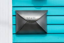 A High Black Metal Postal Box Or Letterbox On A Bright Blue Wooden Clapboard Exterior House Wall. The Vertical Mailbox Is Similar To An Envelope. The Wooden Wall Is Of Narrow Bright Teal Green Board.