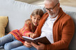 © Pixel-Shot - Little girl with her grandfather reading book at home