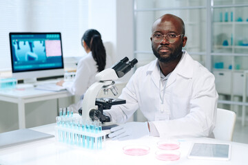 Aufkleber - Young serious scientist in lab coat sitting by workplace in front of microscope and looking at camera during scientific work