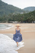 Woman in blue dress and straw hat walk on the beach.
