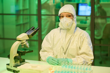 Aufkleber - Young contemporary scientist in biohazard suit sitting by workplace with microscope and group of flasks with liquid substance in laboratory