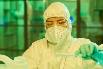 Aufkleber - Young contemporary female scientist in protective coveralls, gloves and respirator pouring blue liquid from flask into other glassware