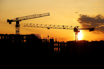 Wall Mural - Silhouettes of construction cranes and workers on scaffolding of unfinished building at sunrise. Housing construction, apartment block in city on dramatic sky background