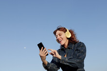 Half Body Shot Of A Woman With Curly Red Hair Smiling With Headphones While Pressing The Screen Of The Cell Phone She Is Holding In Her Hand With A Blue Sky Background