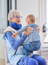 Grandma In Casual Clothes With Glasses And Gray Hair Cooing And Playing With Baby While Sitting On Chair In Dining Room At Home.