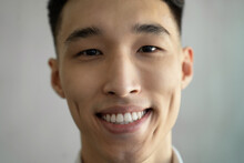 Asian Man With Dimples Smiles Widely Raising Eyebrow In Room On Blurred Background. Portrait Of Young Attractive Guy Looking In Camera Closeup