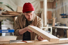 Horizontal Medium Portrait Of Young Caucasian Man Working In Joinery Workshop Checking Wooden Plank Surface Before Polishing It