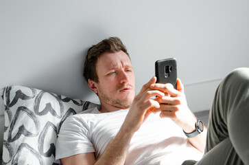 Caucasian attractive young man in white t-shirt woke up chatting, surfing, working on pc and making phone call on bed in bedroom interior. Modern device for remote communication