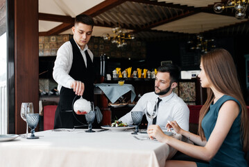 Wall Mural - A young waiter in a stylish apron serves a table with a beautiful couple in an elegant restaurant. Customer service in an elite restaurant and public catering establishment.