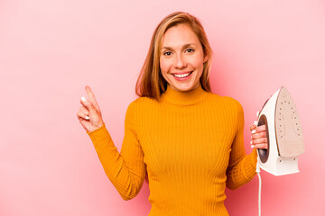 Young caucasian woman holding iron isolated on pink background smiling and pointing aside, showing something at blank space.