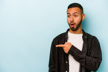 Young Hispanic Man Isolated On Blue Background Points With Thumb Finger Away, Laughing And Carefree.