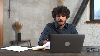 Wall Mural - Hilarious Indian guy using laptop for remote video connection, cheerful multiracial male employee holding virtual meeting, sitting at the desk in modern office, looks at webcam and waving hand