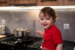 Wesoły chłopiec w czerwonej koszulce w kuchni, z tyłu na kuchence garnek z parującym obiadem.