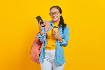 Wall Mural - Portrait of smiling young Asian woman student in denim clothes with backpack holding cup of coffee and using smartphone isolated on yellow background