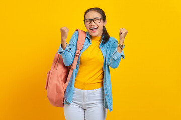 Wall Mural - Cheerful young Asian student in denim outfit with backpack standing doing winning gesture celebrating fist saying yes isolated on yellow background. Education in high school university college concept