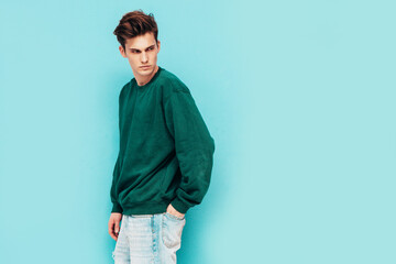 Portrait of handsome confident stylish hipster lambersexual model.Man dressed in green sweater and jeans. Fashion male posing in studio near blue wall