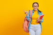 Portrait of smiling young Asian woman college student in denim clothes with backpack holding notebook and pointing finger at copy space isolated on yellow background
