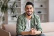 Smiling attractive millennial middle eastern guy blogger with beard looks at camera, sits on sofa in living room