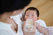 Cropped View Of Asian Mom Looking At Baby Drinking Breast Milk From Bottle. High Angle View Of Lovely Newborn Having Milk With Eyes Close.