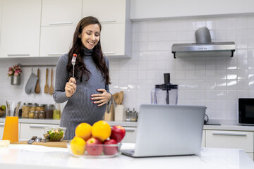 Healthy and cheerful pregnant woman preparing food for herself in her home kitchen. The menu is a vegetable and fruit salad.