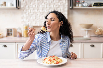 young latin woman eating delicious pasta, enjoying tasty homemade lunch with closed eyes, sitting in