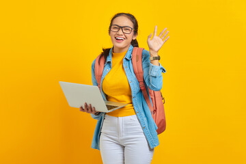 Wall Mural - Portrait of cheerful young Asian woman student in casual clothes with backpack using laptop and gesturing waving hand isolated on yellow background. Education in college university concept