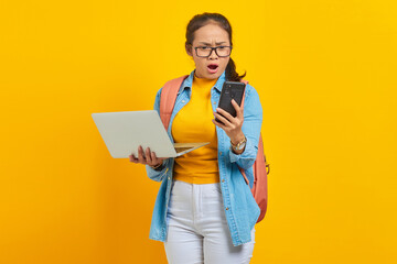 Wall Mural - Shocked young Asian woman student in casual clothes with backpack holding laptop and reading news message on smartphone isolated on yellow background. Education in college university concept