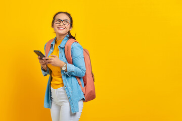 Portrait of smiling young Asian woman student in casual clothes with backpack using mobile phone and looking aside isolated on yellow background. Education in college university concept