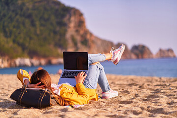 Wall Mural - Millennial freelancer female using mockup laptop with empty screen blank and lying on the sand beach by the sea. Dream office remote work concept