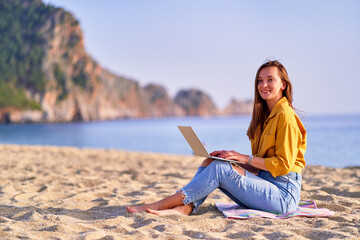 Young happy joyful satisfied millennial freelancer girl using laptop on the beach by sea. Dream office work concept